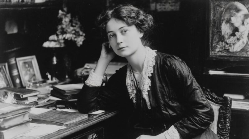 A vintage black-and-white photograph of a woman in early 20th-century attire sitting at a desk filled with books, resting her head on her hand